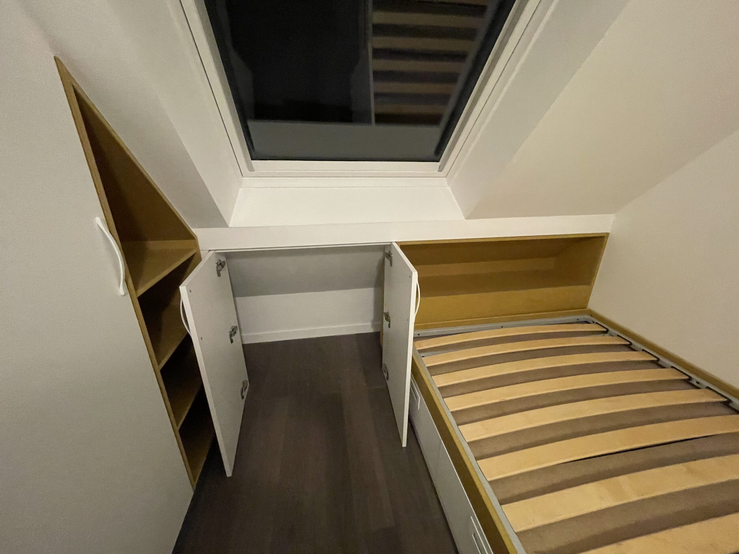 Chambre mansardée avec lit en bois, placards blancs sur mesure sous une fenêtre de toit et parquet sombre.