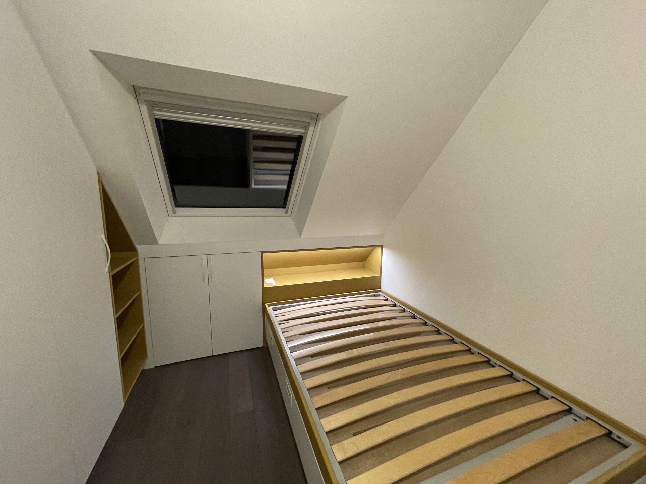 Lit en bois dans une petite chambre mansardée avec placards blancs intégrés et fenêtre de toit.
