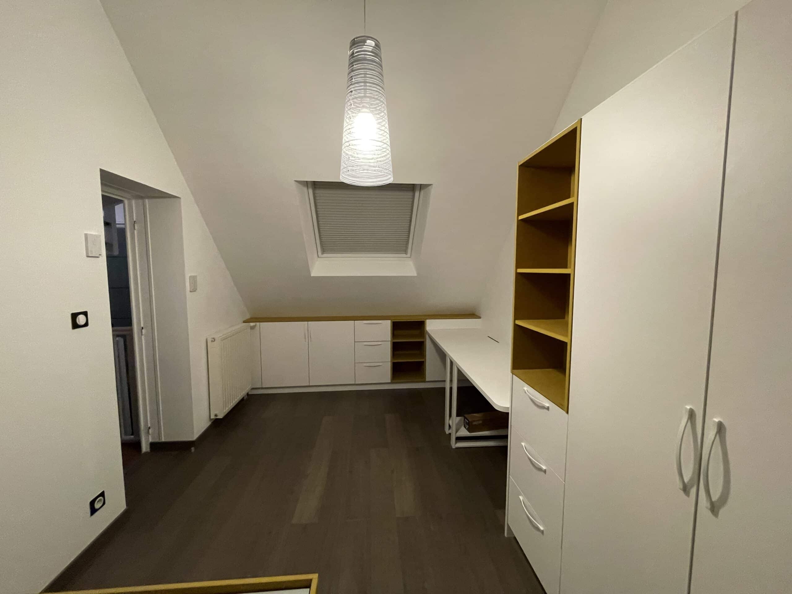 Bureau sous pente moderne avec meubles blancs et bois, étagères jaunes et luminaire design sur parquet sombre.