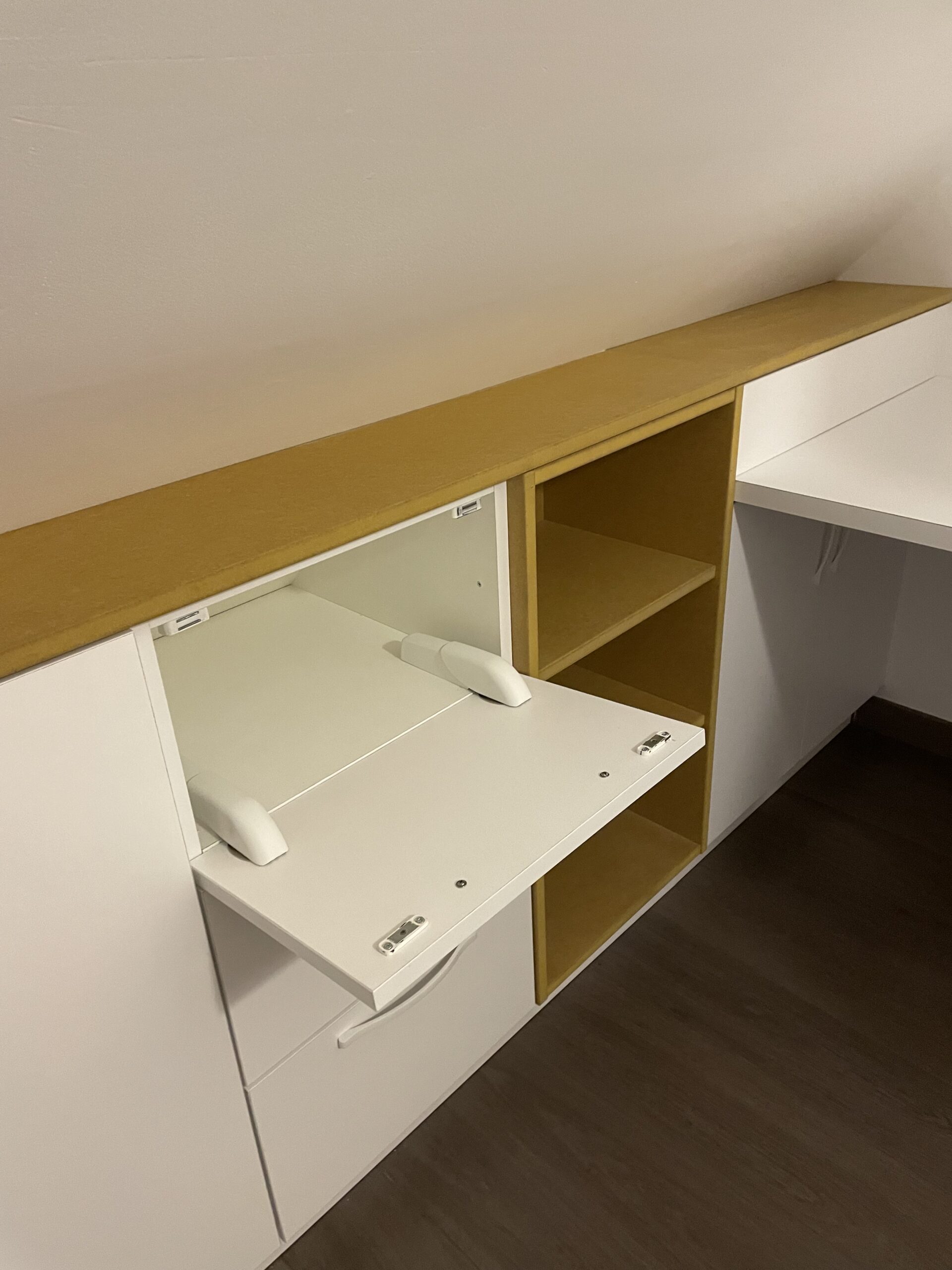 Meuble sur mesure sous pente avec porte abattante blanche ouverte et étagères en bois jaune moutarde.