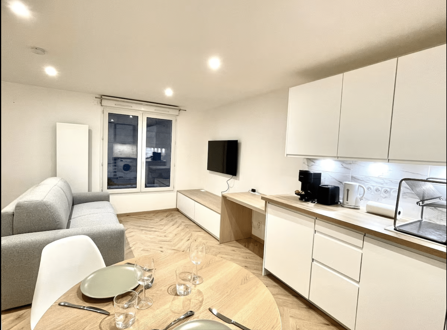 Studio moderne avec cuisine blanche, parquet en chevron, salon avec canapé gris et coin repas dressé.