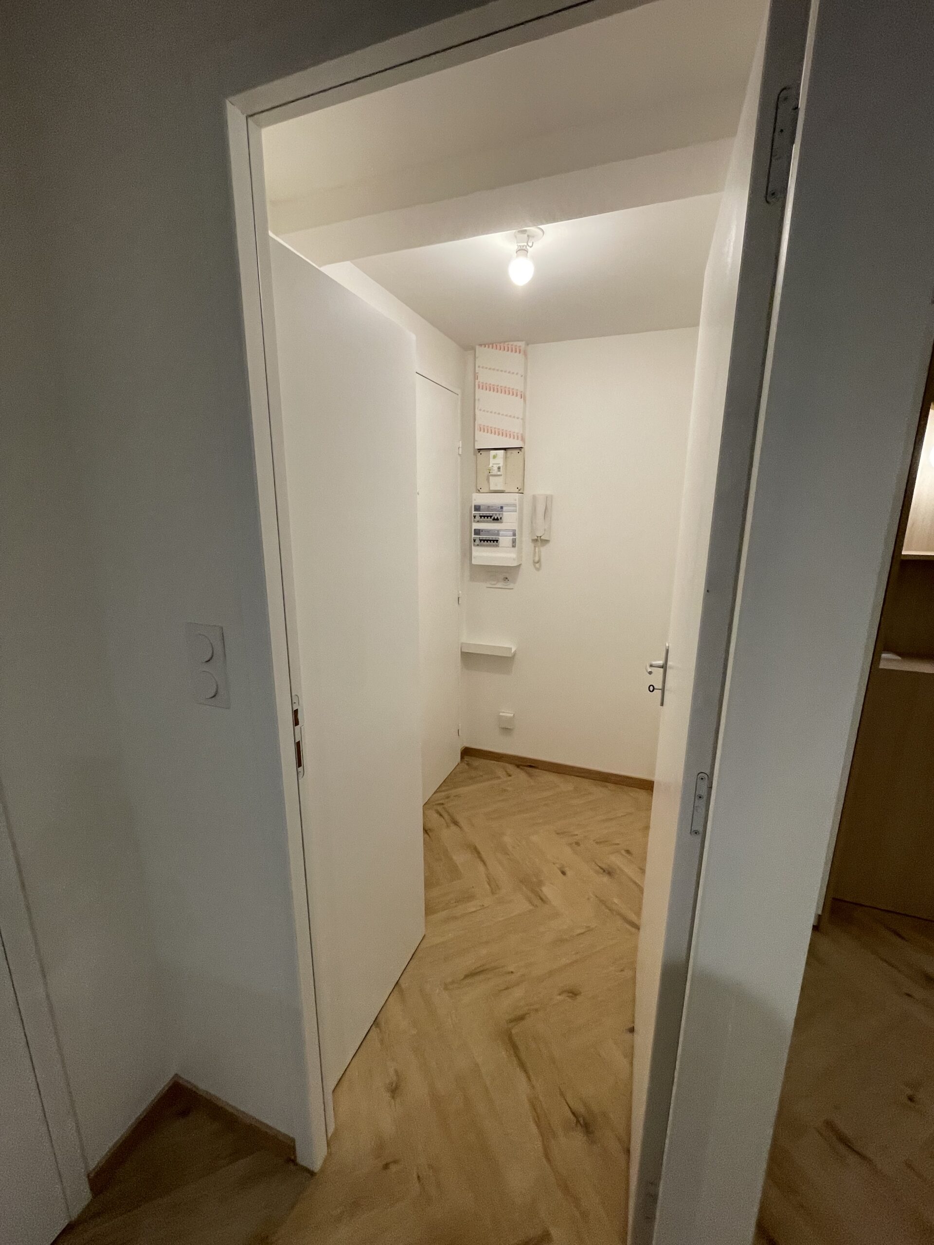 Couloir moderne avec parquet en chevrons, murs blancs, tableau électrique et intercom vus à travers une porte ouverte.
