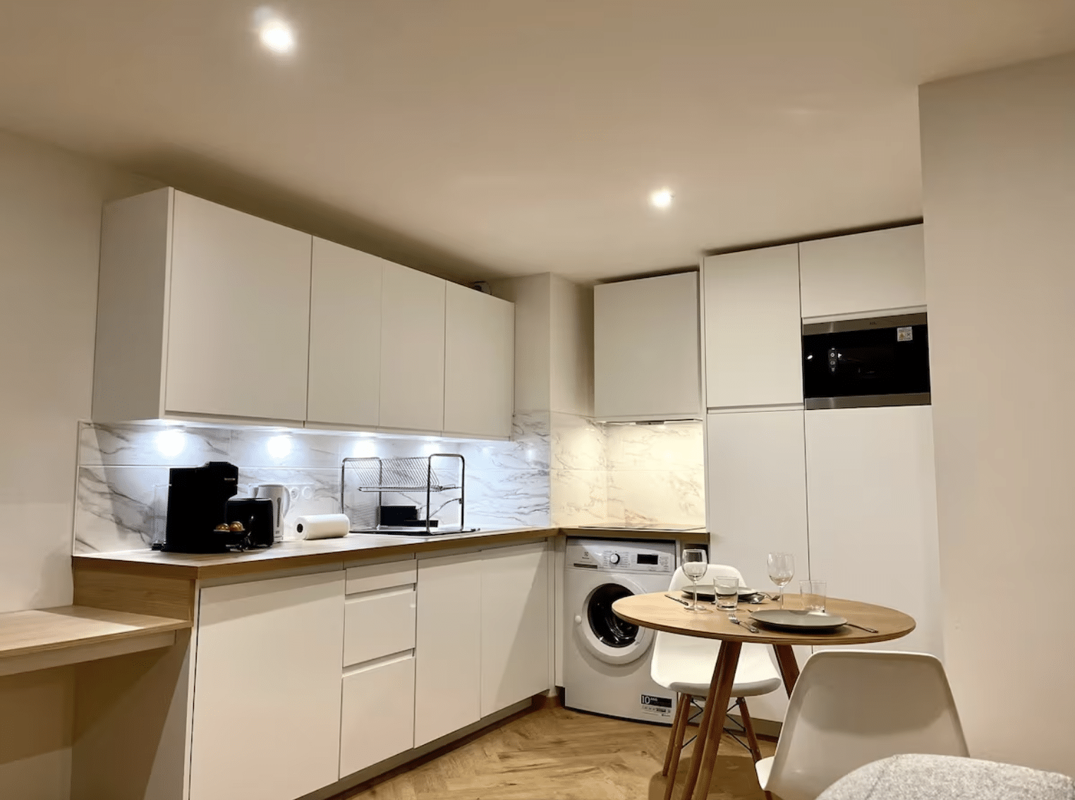 Cuisine moderne blanche avec coin repas, plan de travail bois, lave-linge et crédence effet marbre.