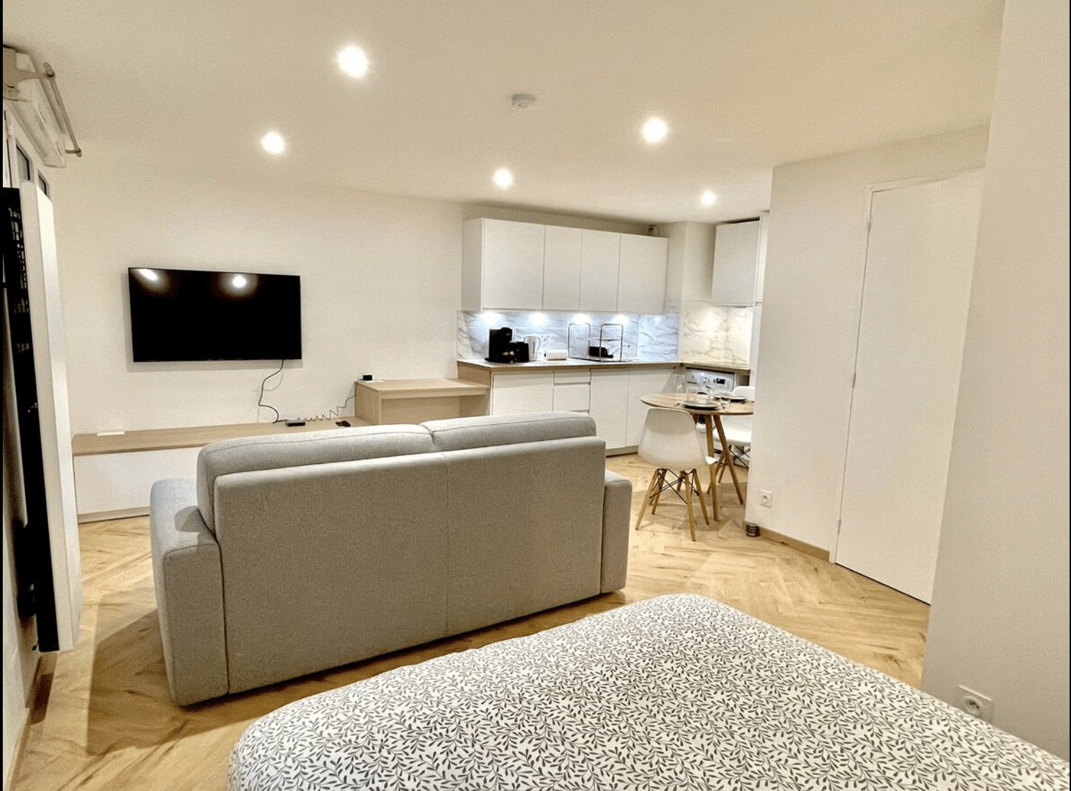 Studio moderne avec canapé gris, TV murale, cuisine ouverte blanche et coin lit dans un espace lumineux et épuré.