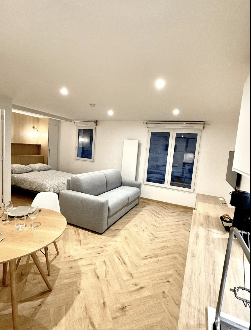 Studio moderne lumineux avec parquet en chevrons, canapé gris, lit double et coin repas.