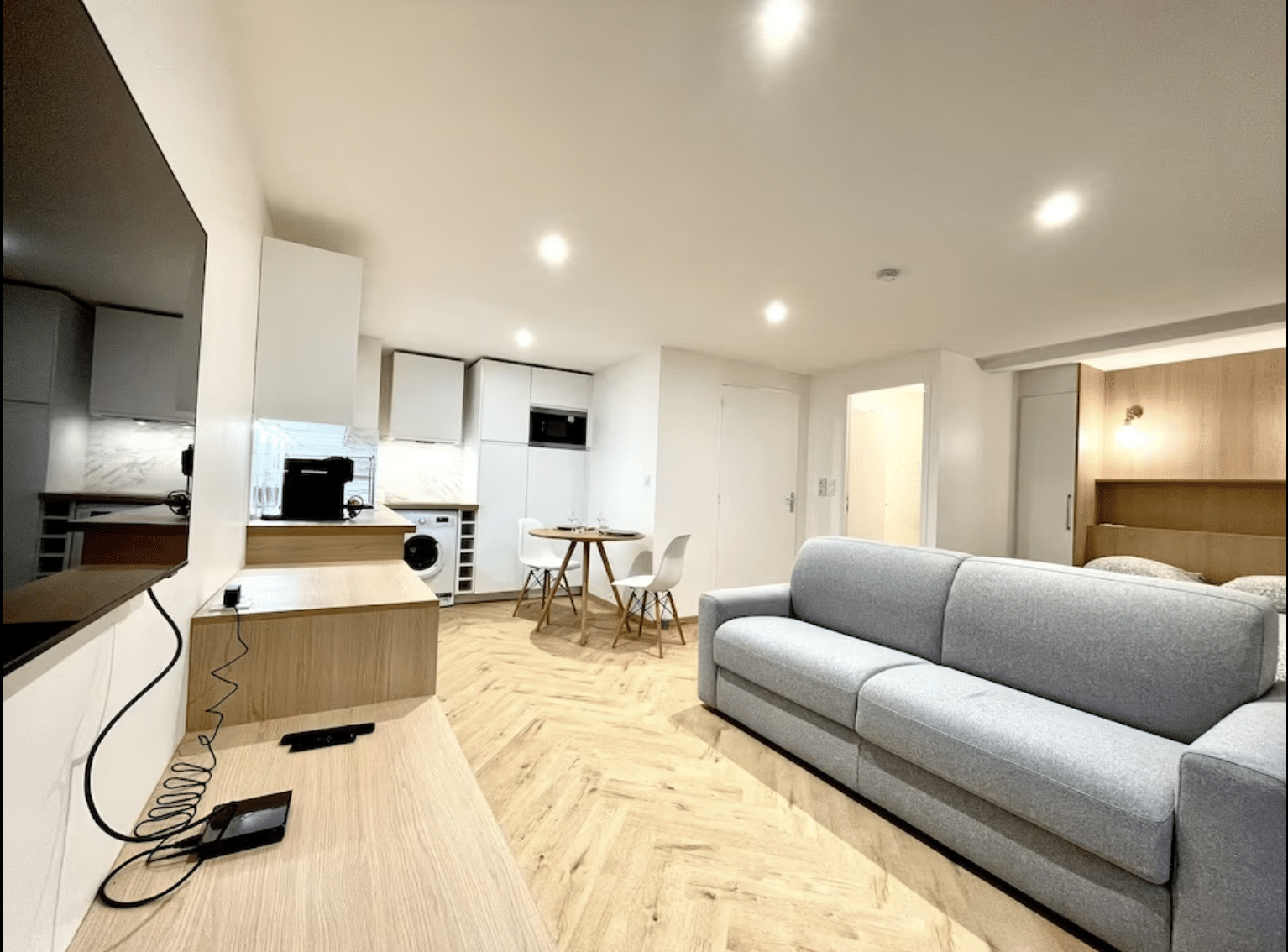 Studio moderne avec canapé gris, parquet en chevrons, kitchenette équipée et coin repas lumineux.