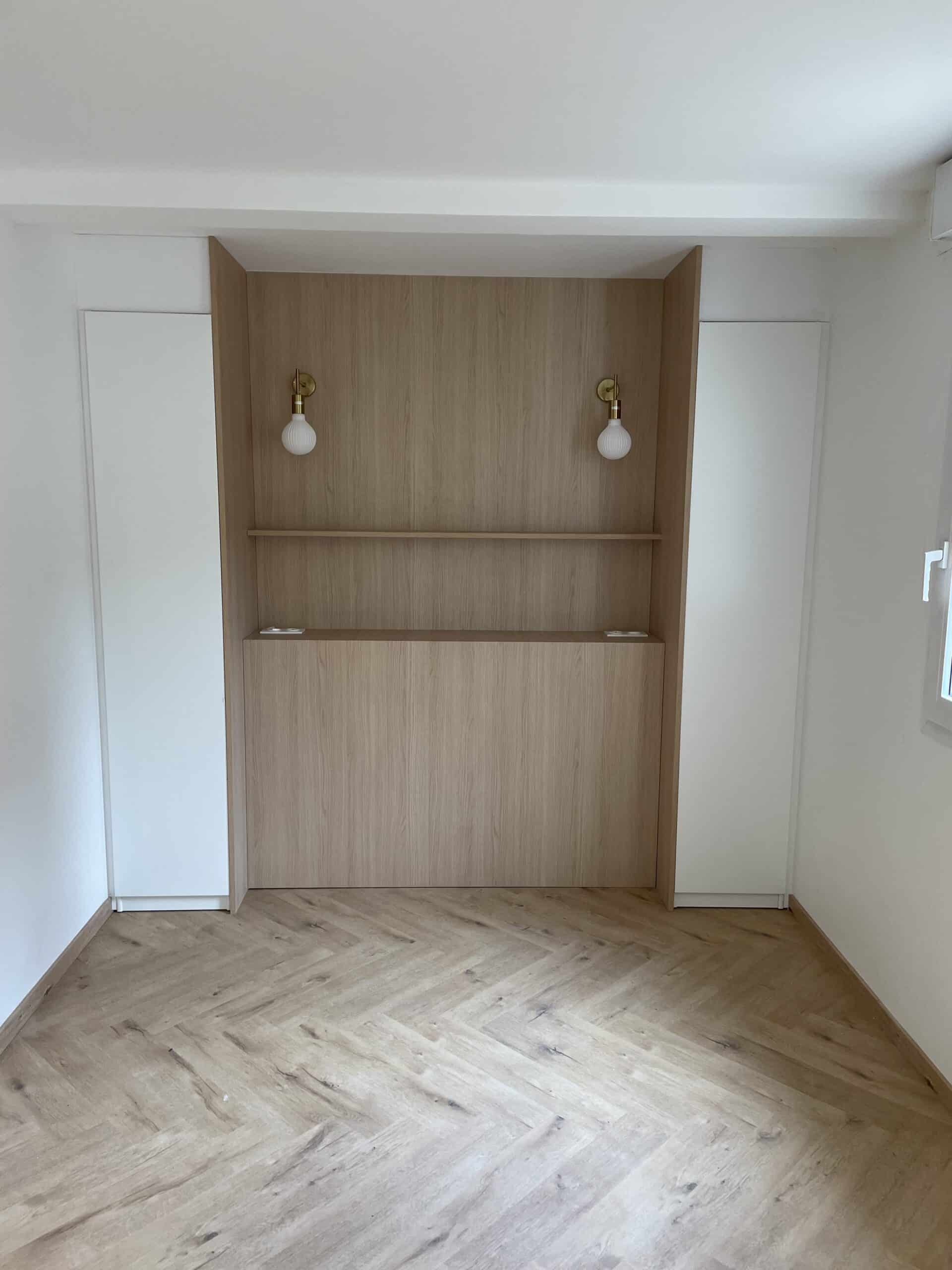 Alcôve de chambre avec tête de lit en bois, appliques en laiton et placards blancs sur parquet en chevrons.