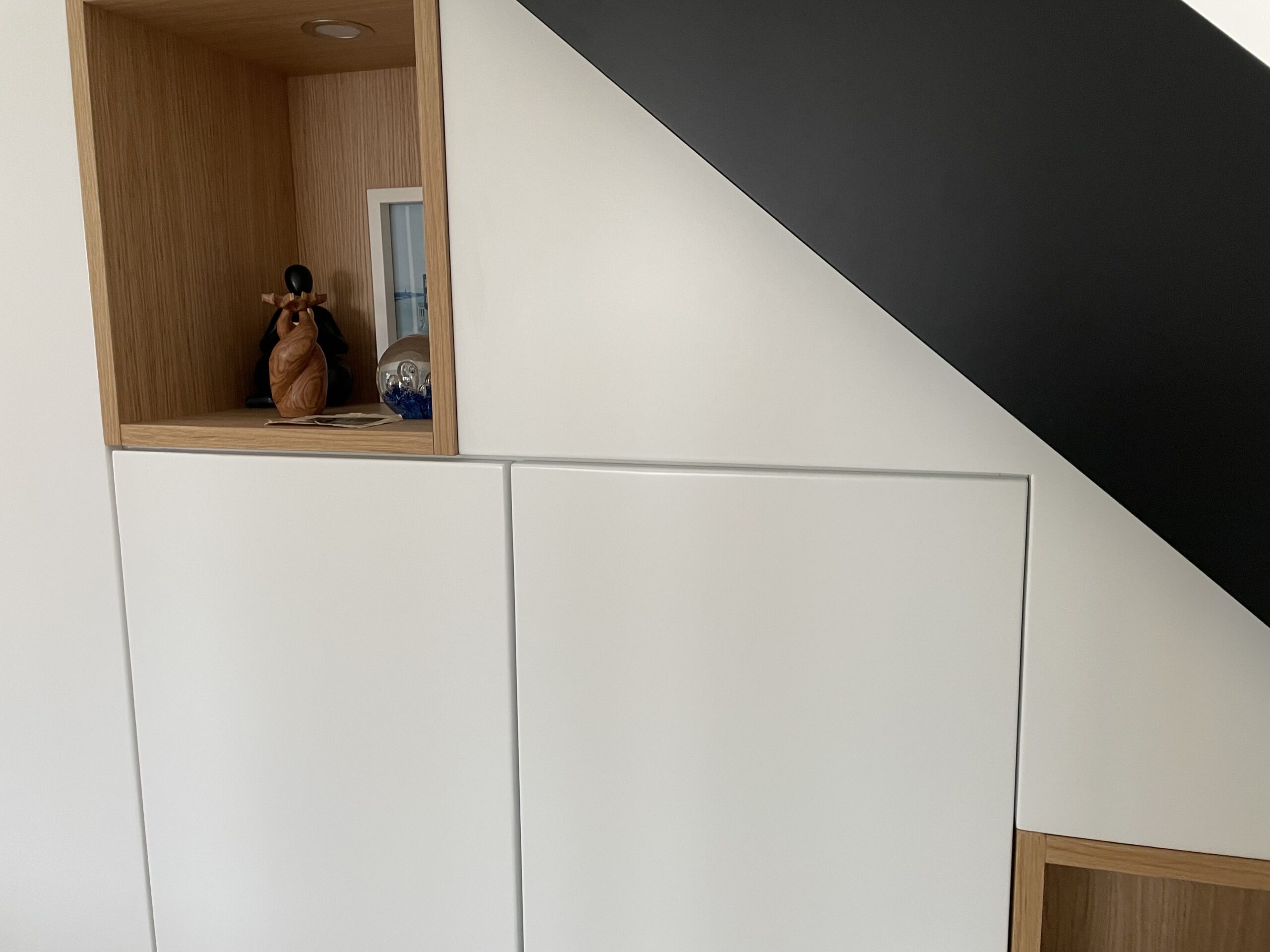 Rangement sous escalier moderne : placards blancs, niche en bois clair et objets déco.
