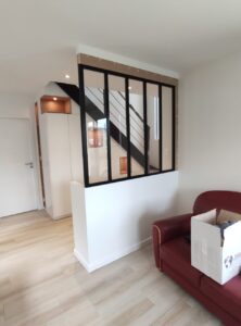 Verrière noire séparant salon et escalier moderne, canapé rouge sur parquet clair.
