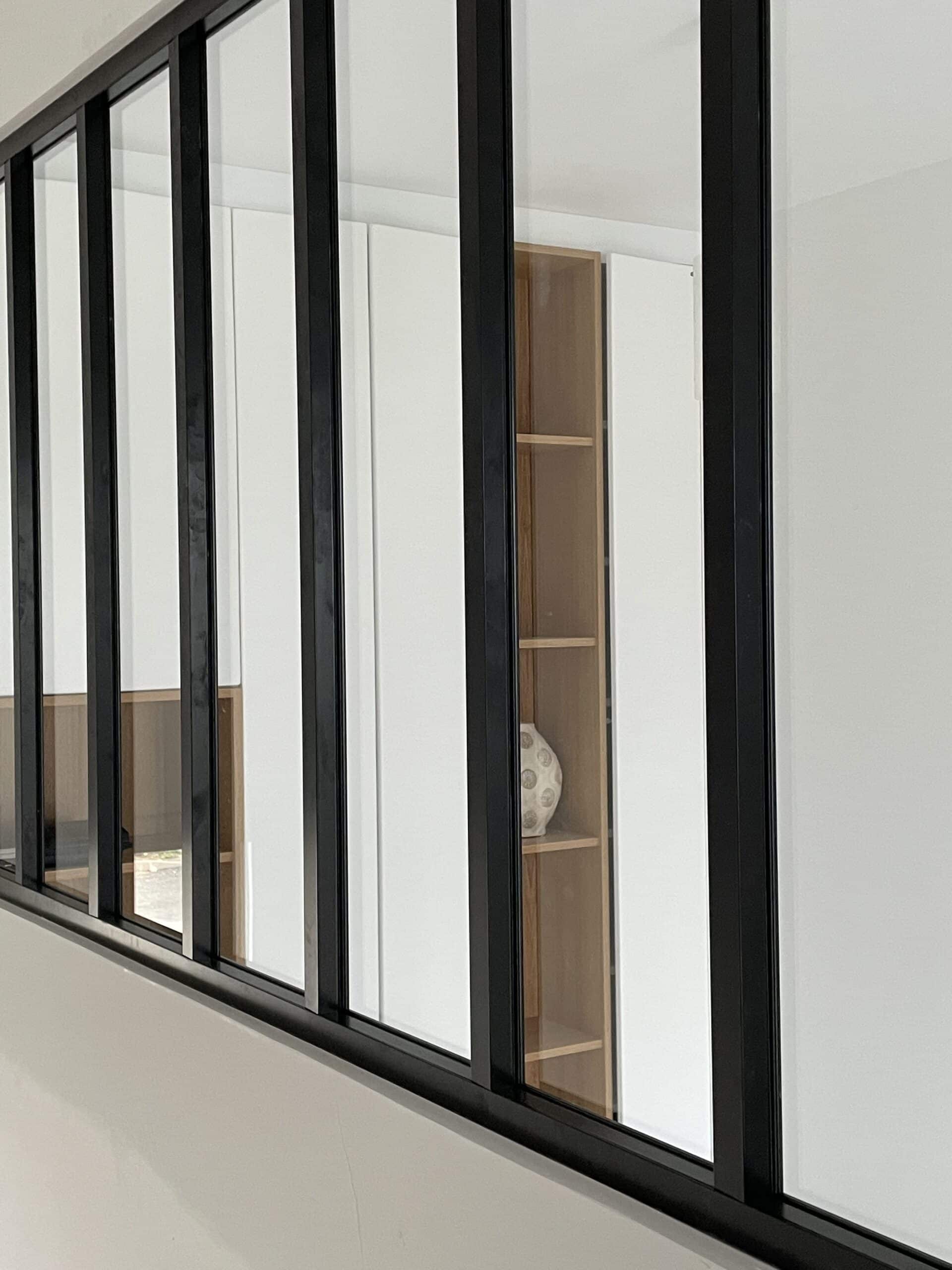 Verrière d'intérieur en métal noir de style industriel, avec vue sur une bibliothèque en bois et murs blancs.